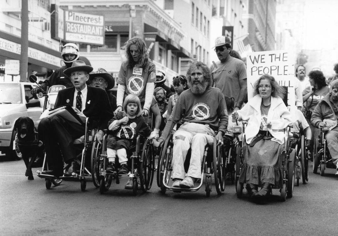 Disability rights leaders in wheelchairs, including Justin Dart and Jennifer  (as a child), protest on the street in San Francisco.