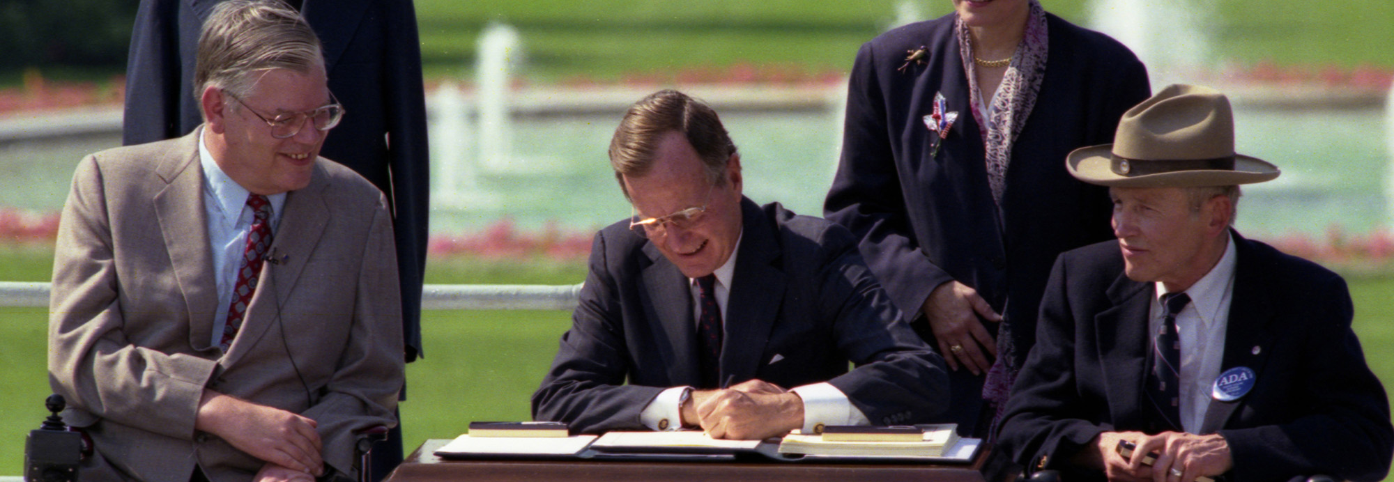President George H. W. Bush signs the Americans with Disabilities Act seated outdoors in front of a fountain with disability activist Justin Dart, Jr. and other onlookers.