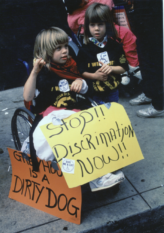 Jennifer Keelan-Chaffins (as a child) in a wheelchair and her sister Kailee Keelan (as a child) protest on the streets of Atlanta holding banners and wearing stickers. 