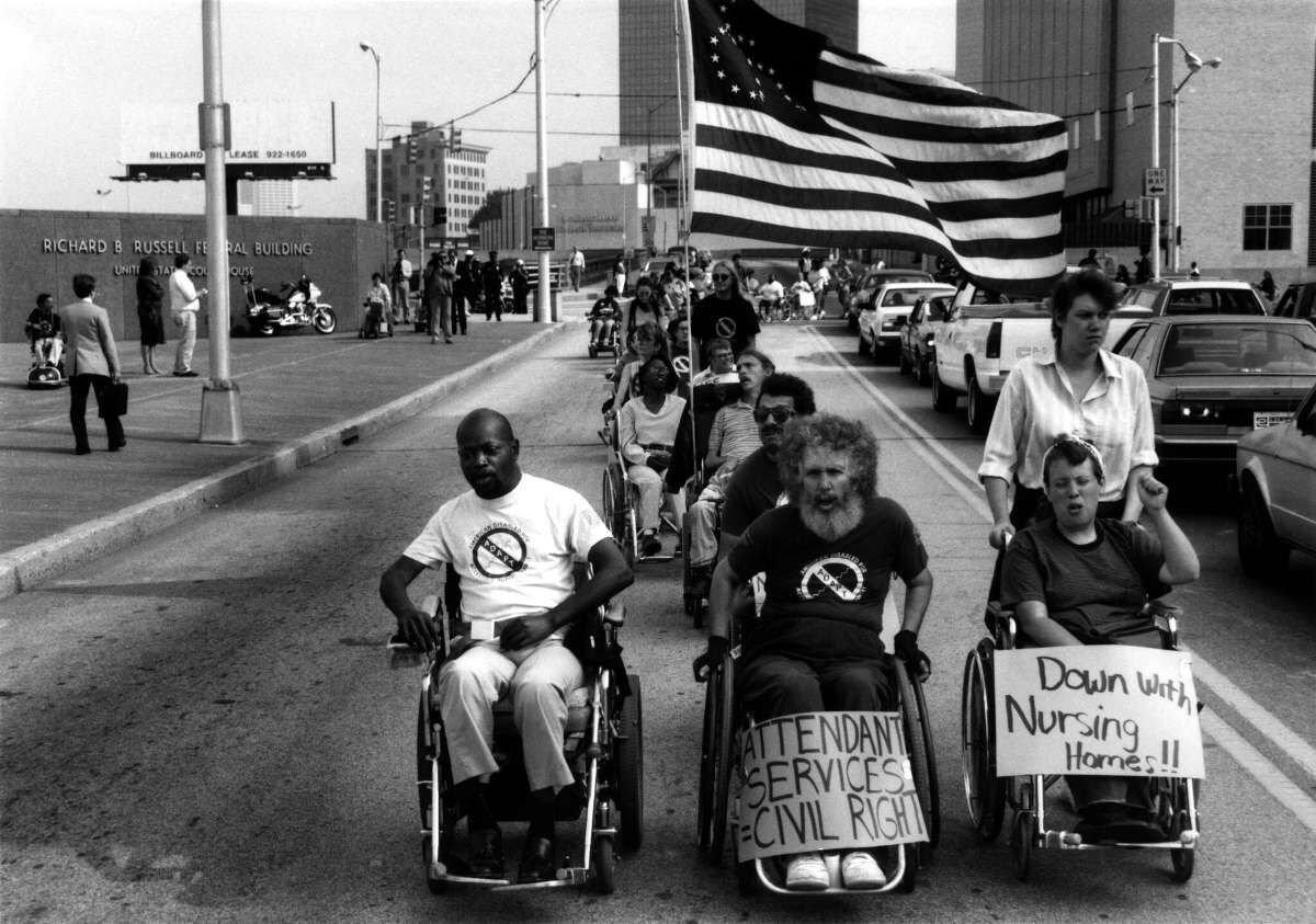 Disability rights activists protest on the streets with signs and a flag.